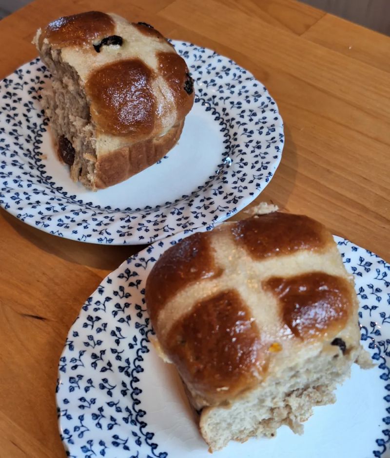 Freshly baked hot cross buns on patterned plates, showing their soft interior and glossy glazed tops.