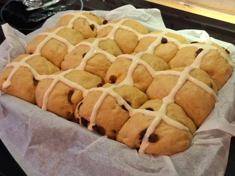 Homemade hot cross buns with raisins and traditional cross topping arranged in a lined baking tin before baking.