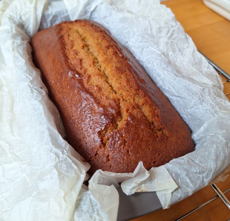 Homemade ginger loaf cake cooling in a lined loaf tin, showing its golden-brown crust and characteristic crack along the top.