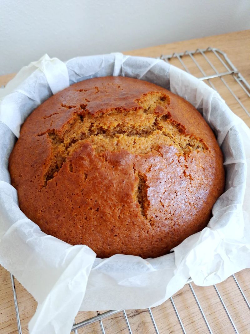 Homemade round ginger cake cooling in a lined cake tin, with a golden-brown crust and cracked top.