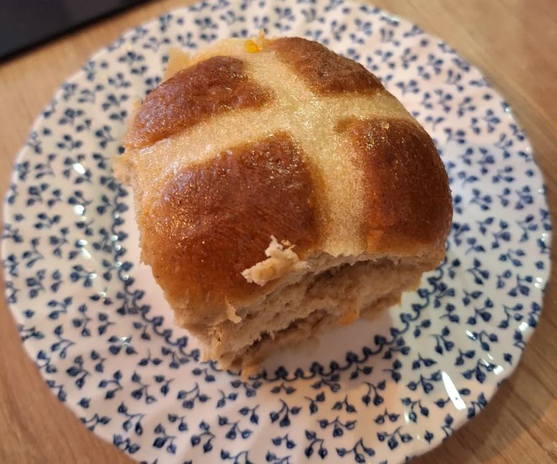 Close-up of a homemade hot cross bun with a glossy glazed top and soft, fluffy interior on a patterned plate.