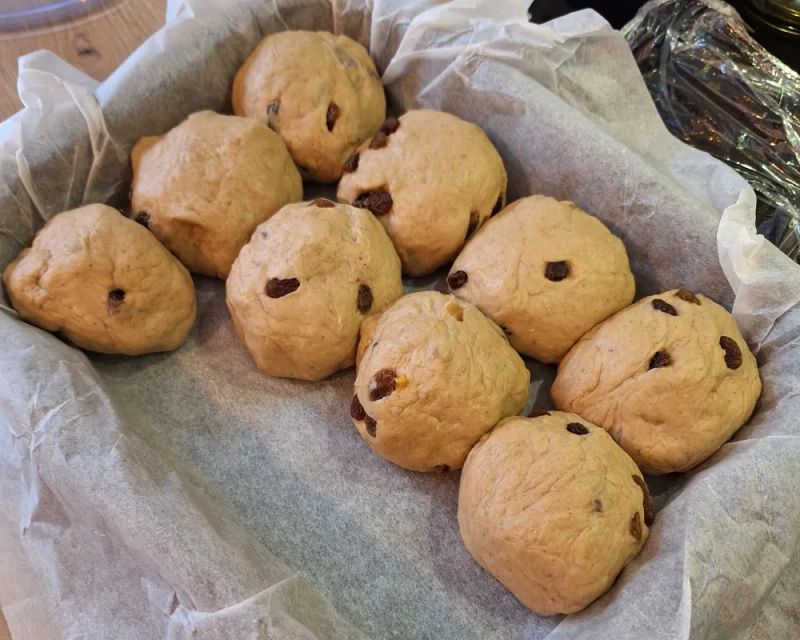 Hot cross bun dough balls with raisins arranged in a parchment-lined baking tin before the crosses are added.